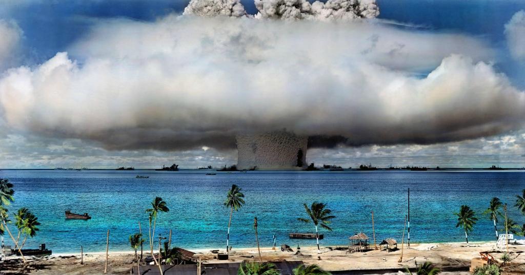 Colorized image of a mushroom cloud with blue sea, palm trees, and beach in the foreground. 