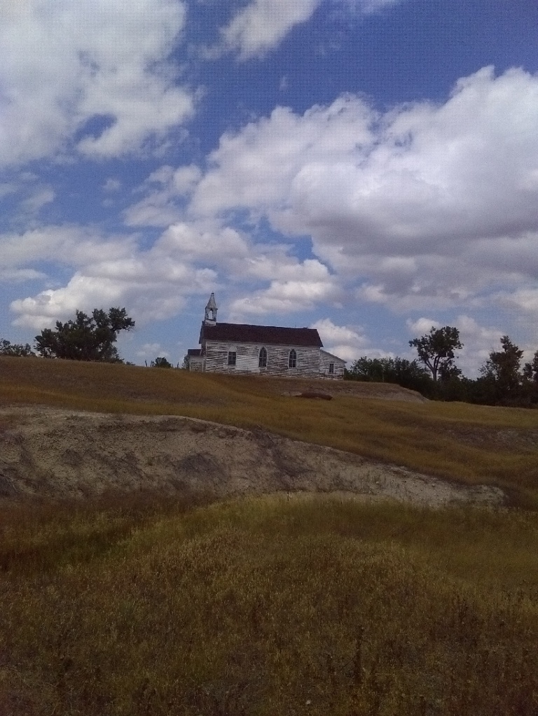 An old, abandoned church with peeling white paint on top of a hill with the blue sky and white clouds behind it. 